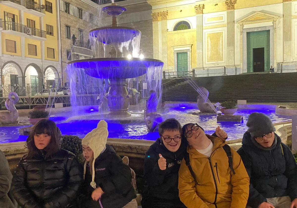 Un gruppo seduto sul bordo di una fontana illuminata di blu, in una piazza di sera.