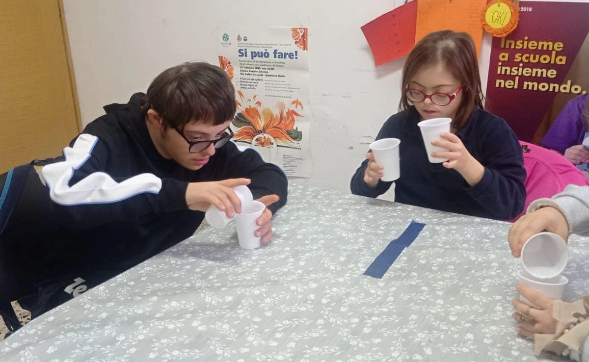 Alcuni ragazzi travasano dell'acqua da un bicchiere di plastica a un altro, seduti a un tavolo.