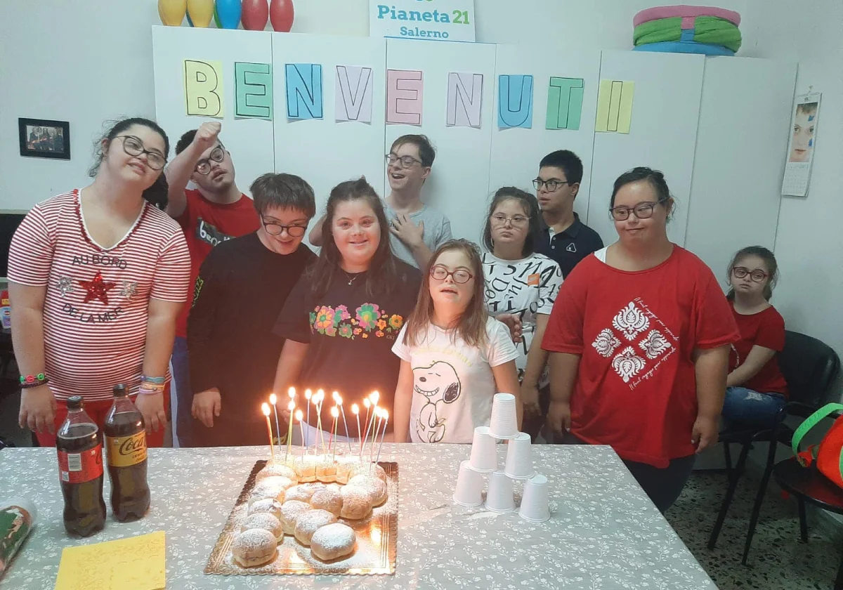 Un gruppo di ragazzi e ragazze in posa dietro a una torta con le candeline accese, sotto uno striscione con la scritta "Benvenuti".
