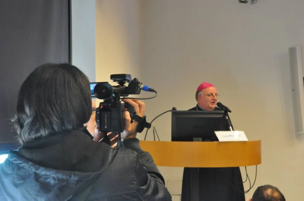 Un relatore in abito talare parla al microfono da un podio durante un convegno, ripreso in primo piano da un'operatrice con una videocamera.