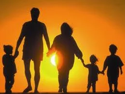 Sagoma in controluce di una famiglia — due adulti e tre bambini — che cammina tenendosi per mano al tramonto.