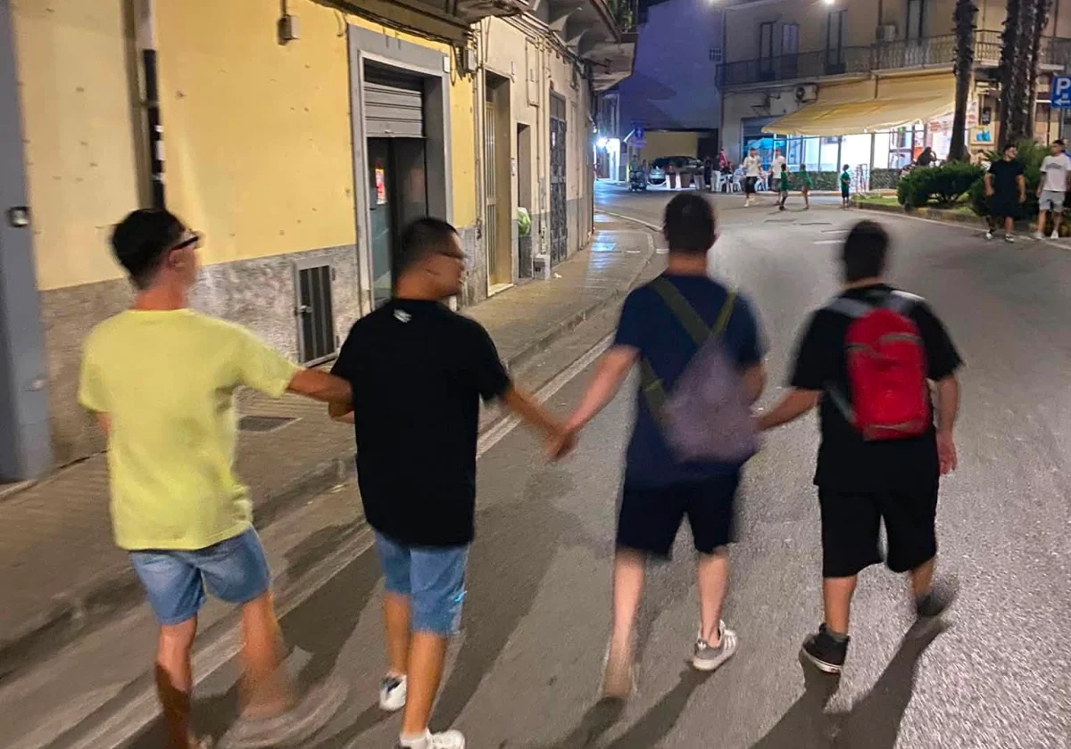 Quattro ragazzi ripresi di spalle mentre camminano per una strada, mano nella mano, di sera.